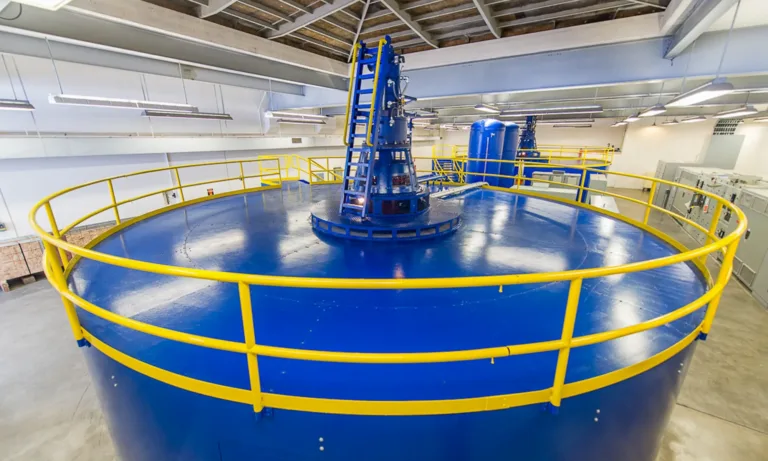 Large blue circular industrial machine with yellow safety railings and a central laddered mechanism inside a brightly lit plant room.