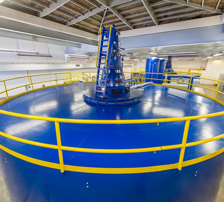 Large blue circular industrial machine with yellow safety railings and a central laddered mechanism inside a brightly lit plant room.