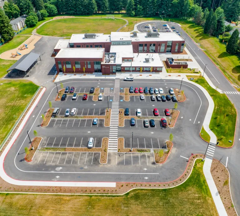 Aerial view of a modern brick school with a large parking lot featuring landscaped islands and crosswalks, a playground and covered area to the left, a looped drop-off drive to the right, and surrounding lawns and trees.