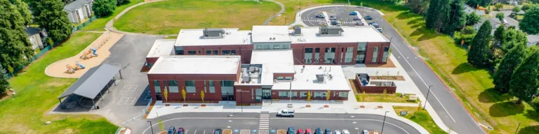 Aerial view of a modern low-rise brick school campus with white roofs, adjacent parking lots and a drop-off loop, a playground and covered sport courts to the left, a large grassy field behind, and tree-lined suburban streets surrounding it.