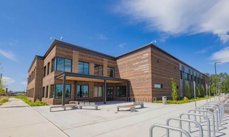 Exterior of Clark College’s Advanced Manufacturing Center (AMC), a modern two-story brick building with large windows, a covered entrance, benches, and bike racks under a clear blue sky.