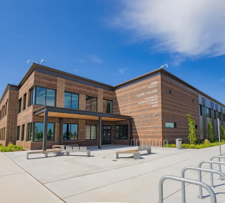 Exterior of Clark College’s Advanced Manufacturing Center (AMC), a modern two-story brick building with large windows, a covered entrance, benches, and bike racks under a clear blue sky.