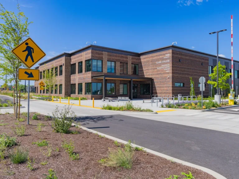 Two-story brick building labeled Clark College Advanced Manufacturing Center on a sunny day, featuring large windows, bike racks, and landscaped sidewalks along a campus road with pedestrian crossing signs.