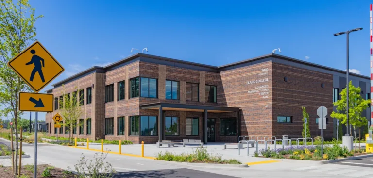 Two-story brick building labeled Clark College Advanced Manufacturing Center on a sunny day, featuring large windows, bike racks, and landscaped sidewalks along a campus road with pedestrian crossing signs.
