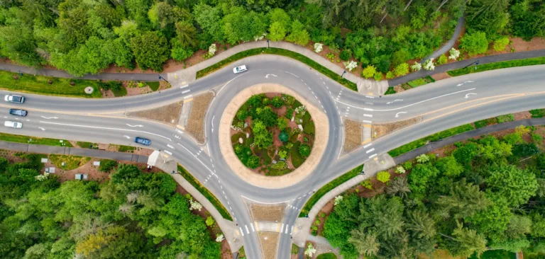 Aerial view of a three-arm roundabout with a landscaped central island, crosswalks, and light traffic, surrounded by dense green trees and nearby homes.