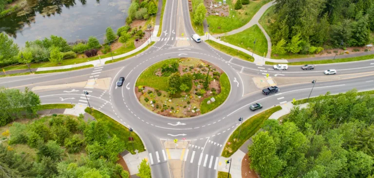 Aerial view of a multi-lane roundabout in a green, wooded area, with cars circulating, crosswalks and yield markings, and a landscaped central island near a small pond.