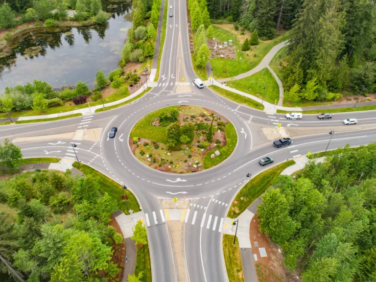 Aerial view of a landscaped multi-lane roundabout with cars and yield markings, crosswalks and walking paths around it, bordered by trees and a small pond.