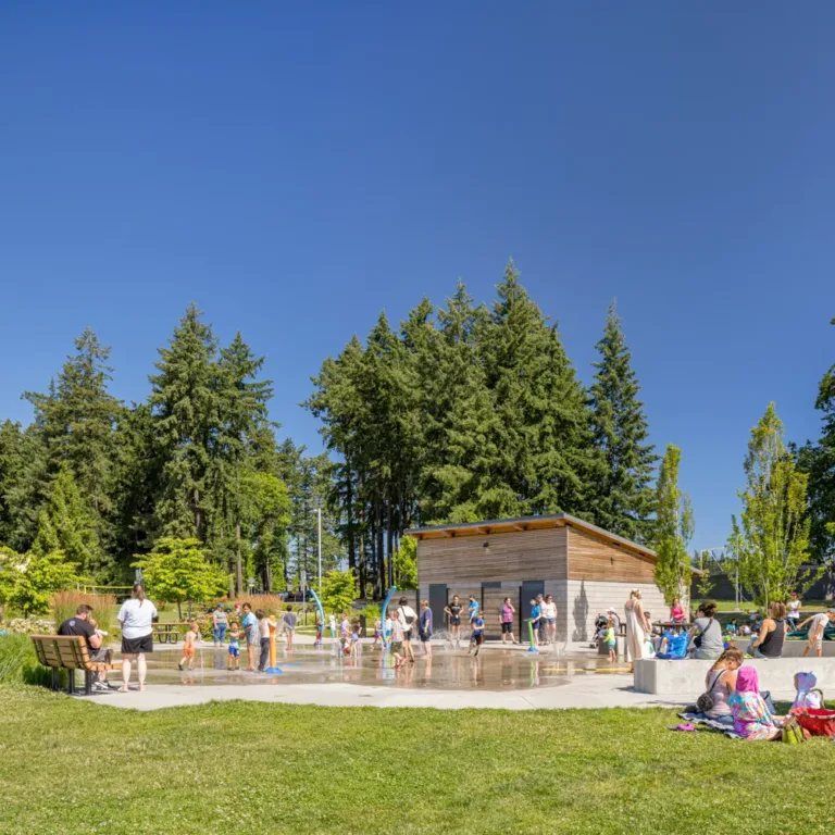 Families gather around a splash pad in a sunny park, with children playing in water jets near a small wood building, benches, and lawns beneath tall evergreen trees and a clear blue sky.