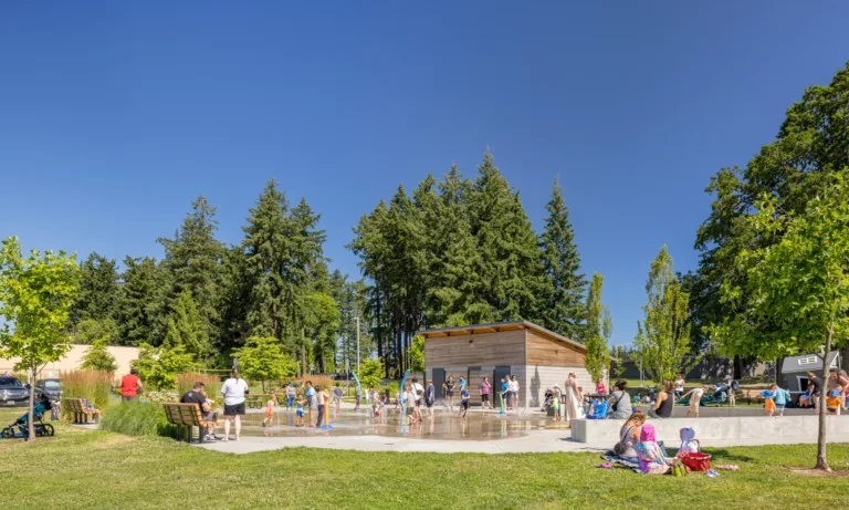 Families and children enjoy a sunny day at a park splash pad, with benches, a small wooden building, and tall evergreen trees under a clear blue sky.