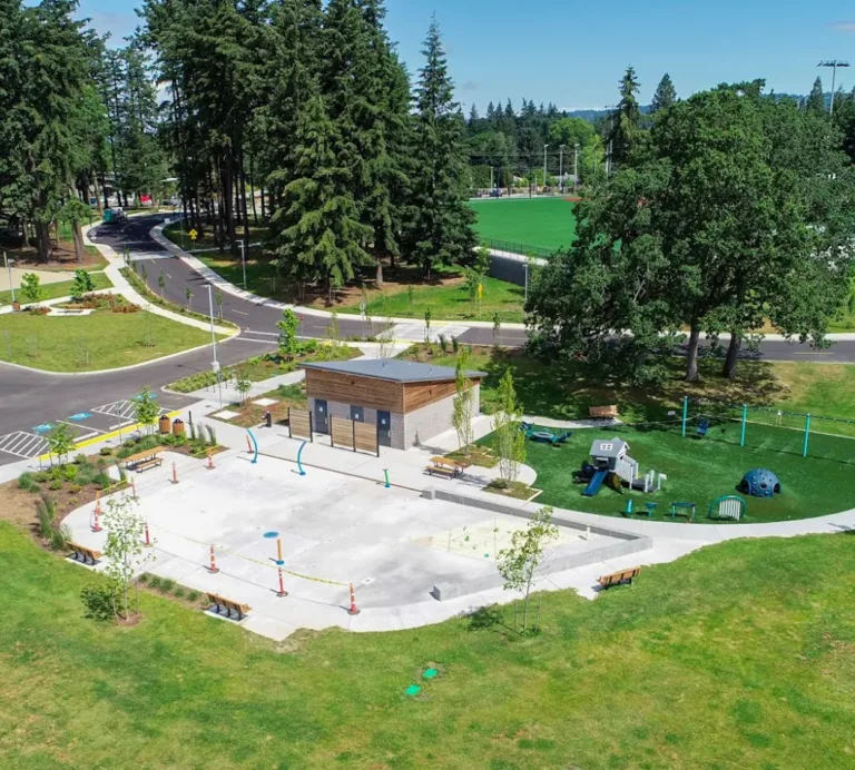 Aerial view of a community park featuring a central splash pad and restroom building, a nearby playground, walking paths and benches, parking areas, and sports fields framed by tall evergreen trees.
