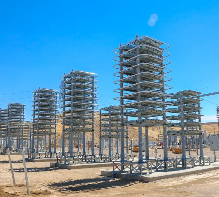 Rows of tall, stacked insulator towers and metal frames at an electrical substation in a desert landscape, with power lines, workers, and equipment under a clear blue sky.