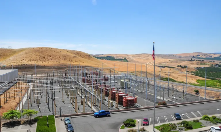 An electrical substation with grids of metal pylons and transformers beside a parking lot, an American flag, and dry rolling hills under a clear blue sky.