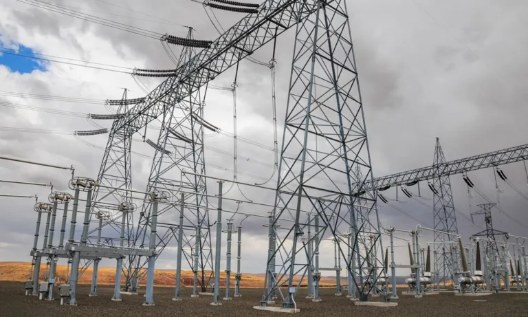 High-voltage electrical substation with lattice towers, insulators, and overhead power lines in an open, arid landscape under cloudy skies.