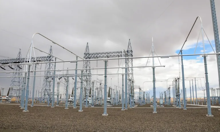 High‑voltage electrical substation with steel towers, insulators, and busbars arranged across a gravel yard under a cloudy sky.