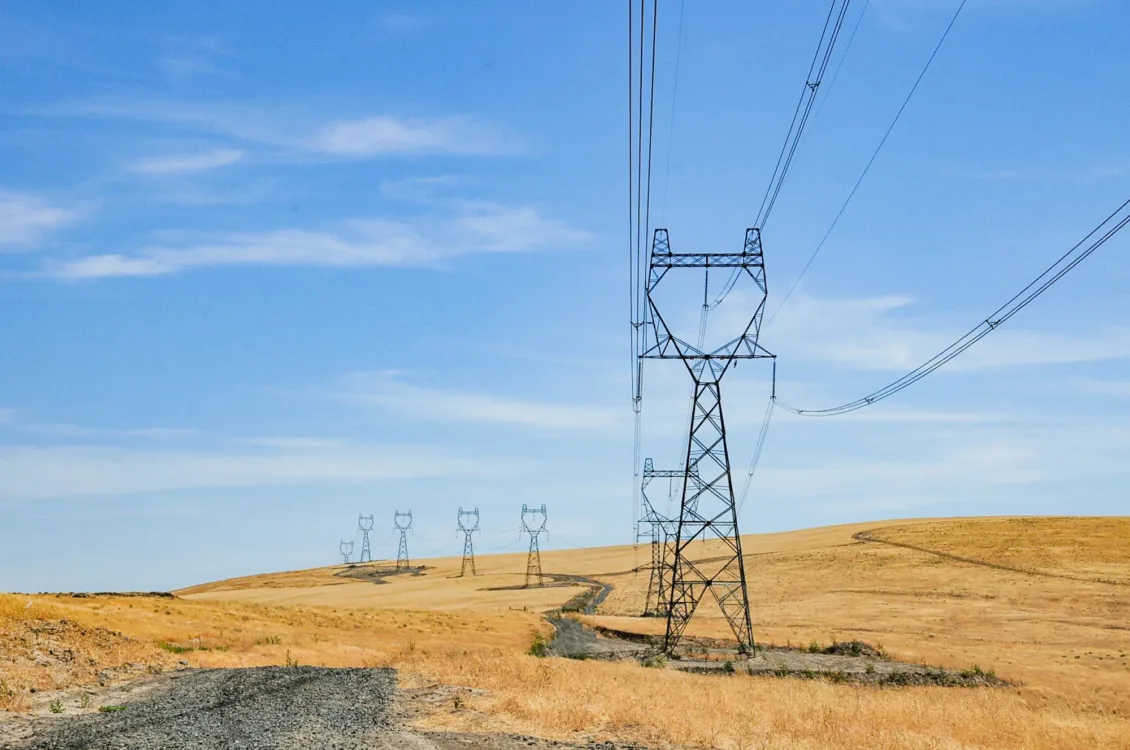 High-voltage transmission towers and power lines stretch across rolling golden grassland with a winding dirt road under a clear blue sky.