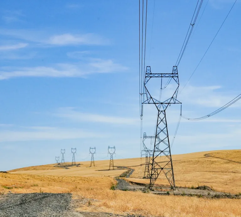 High-voltage transmission towers and power lines stretch across dry, rolling grasslands beside a winding dirt road under a clear blue sky.