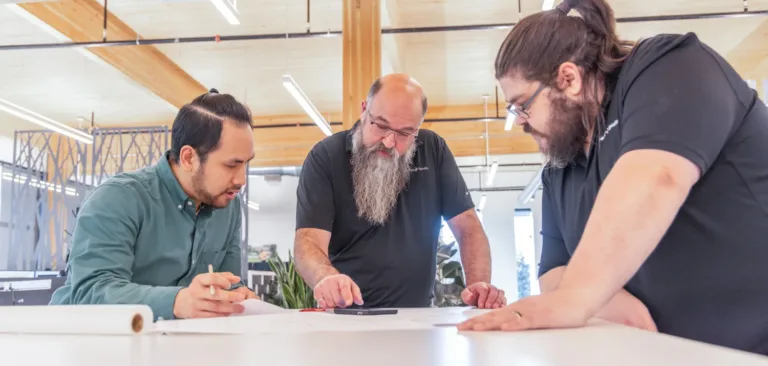 MacKay Sposito team members collaborate over design plans at a large table in a modern office with exposed wood beams and bright industrial lighting.