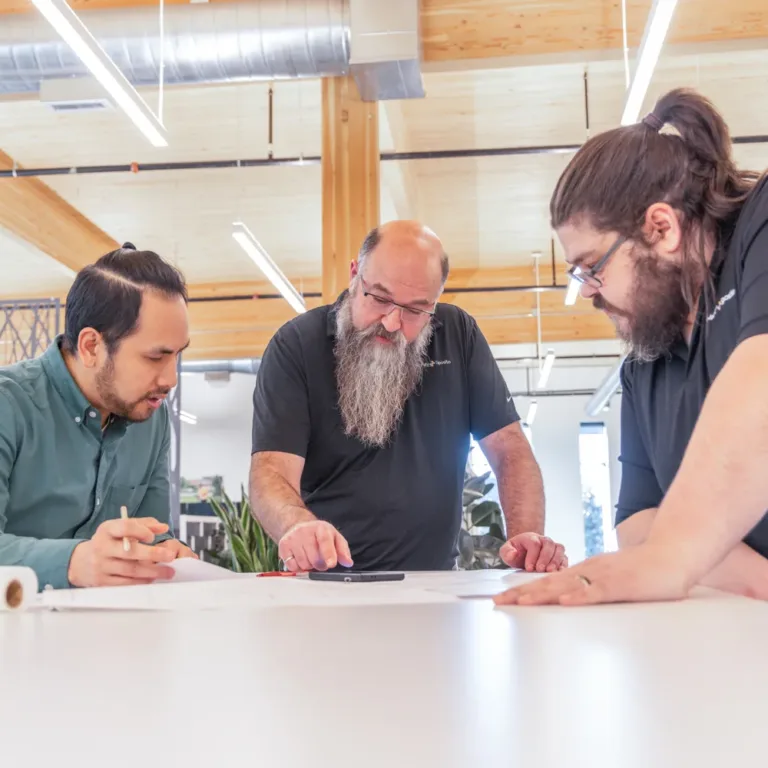 MacKay Sposito team members collaborate over design plans at a large table in a modern office with exposed wood beams and bright industrial lighting.