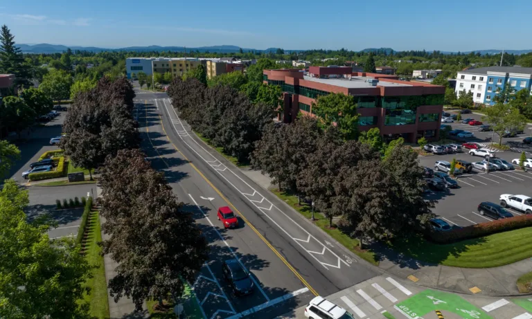 Aerial view of a tree-lined urban street with green-painted bike lanes and a crosswalk, cars traveling past a red brick office building and parking lots under a clear blue sky.