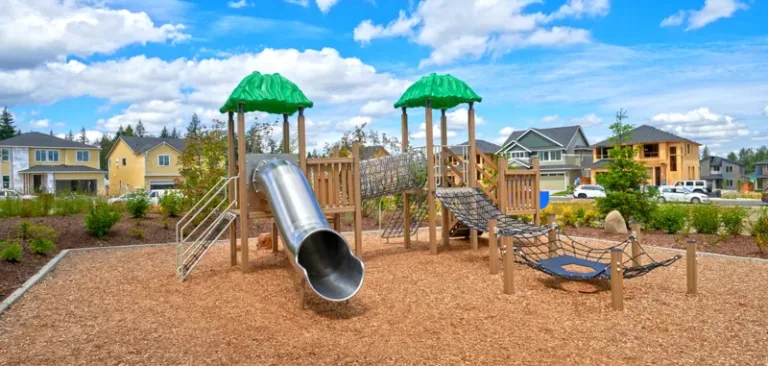 Neighborhood playground with a metal tube slide, rope bridges, and net climbers on a wood-chip surface, featuring green leaf-like canopy tops and houses in the background under a bright blue sky.