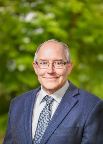 Headshot of Dan Overton smiling, wearing glasses and a blue suit with a patterned tie against a blurred green outdoor background.