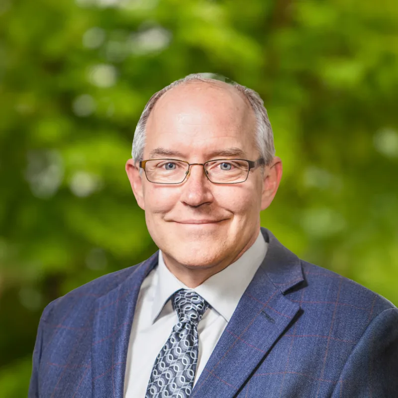 Headshot of Dan Overton smiling, wearing glasses and a blue suit with a patterned tie against a blurred green outdoor background.