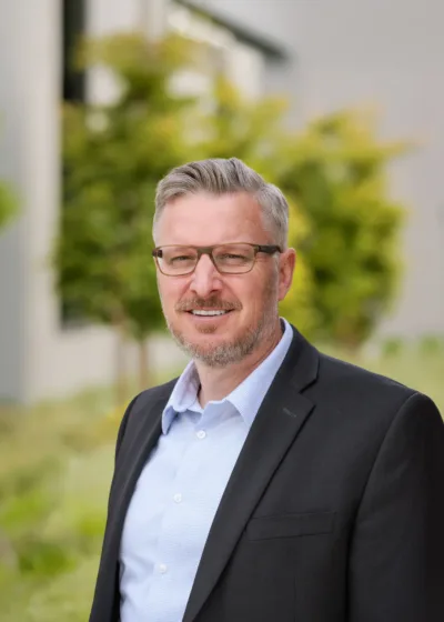 Headshot of Derrick Smith, a person with short gray hair, rectangular glasses, and a light beard, wearing a black blazer over a light blue shirt, smiling outdoors with greenery and a blurred building in the background.