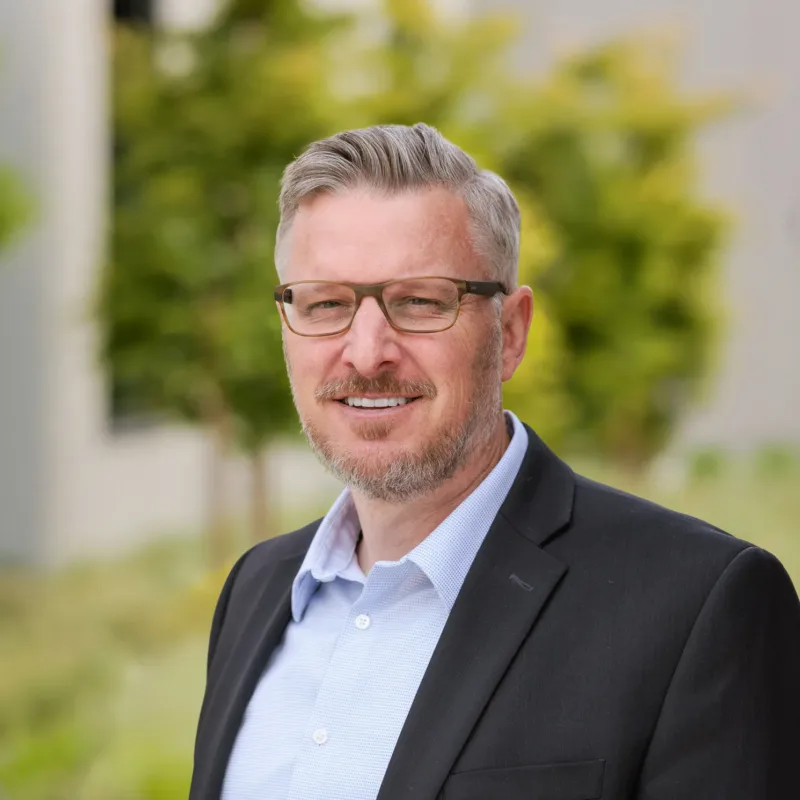 Headshot of Derrick Smith, a person with short gray hair, rectangular glasses, and a light beard, wearing a black blazer over a light blue shirt, smiling outdoors with greenery and a blurred building in the background.