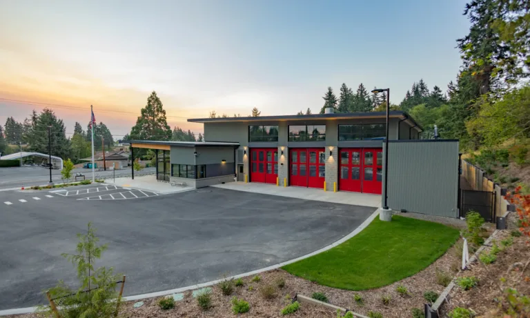 Modern fire station with four bright red bay doors and gray siding, fronted by a wide paved apron and landscaped grounds at dusk, with trees and an American flag in the background.
