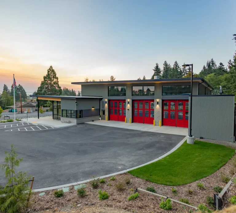 Modern fire station with four bright red bay doors and gray siding, fronted by a wide paved apron and landscaped grounds at dusk, with trees and an American flag in the background.