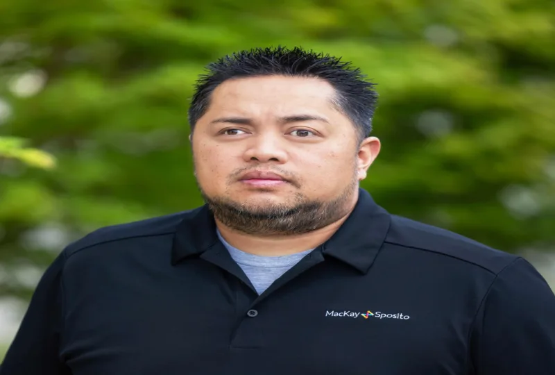 Headshot of Edward Castro, wearing a black MacKay Sposito polo with short spiky hair and a trimmed beard, looking ahead with a neutral expression against a blurred green background.