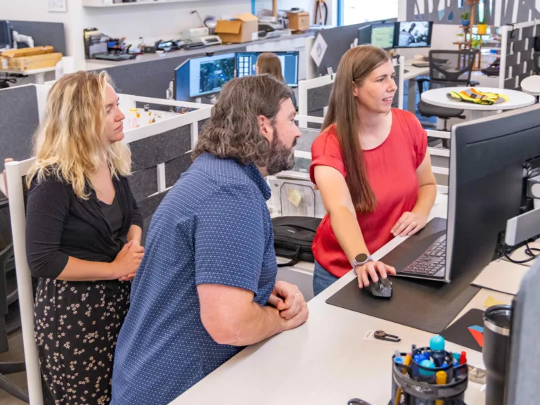 Three coworkers in an open-plan office collaborate at a workstation, with a woman in a red top pointing at a large monitor while two colleagues look on.