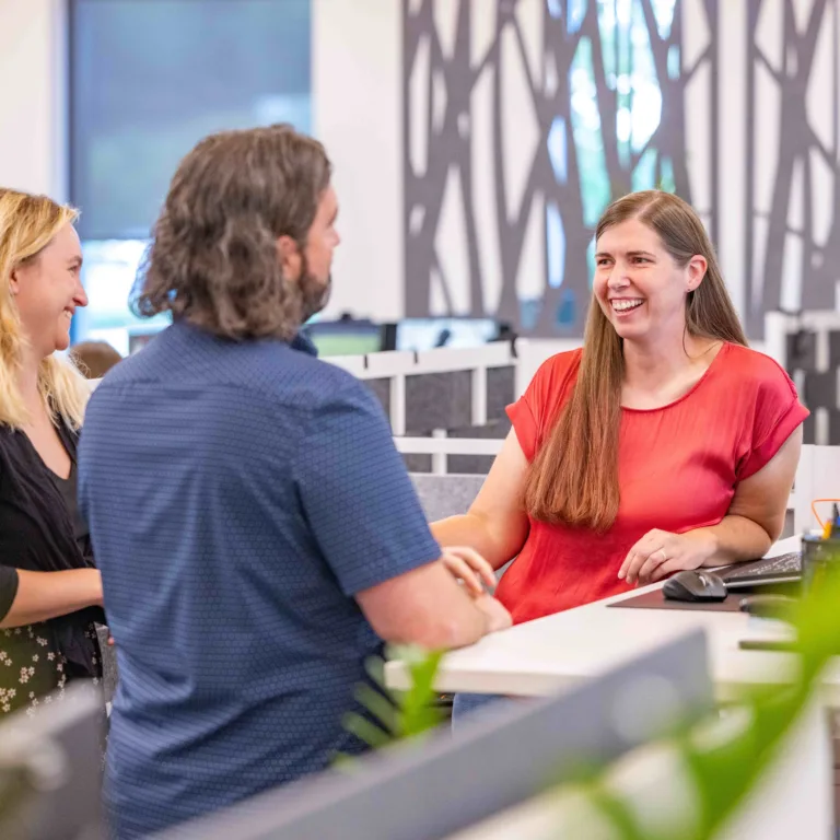 Three colleagues smile and chat at a workstation in a modern open-plan office, with one seated at a computer and two standing across the desk.