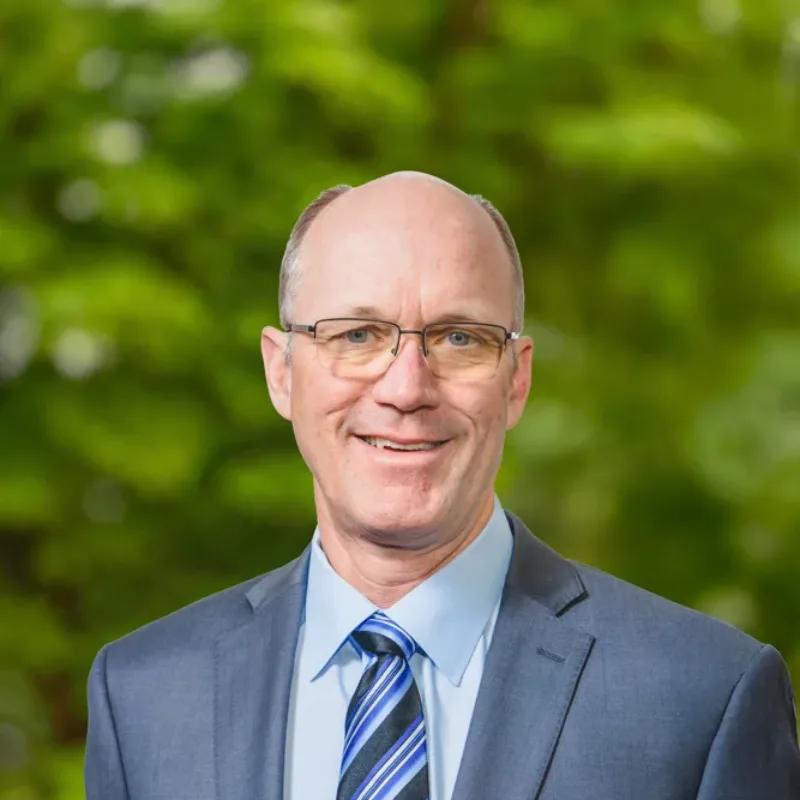 Headshot of Erik Nelson, a smiling man with glasses in a blue suit and striped tie, posed outdoors against a blurred green background.