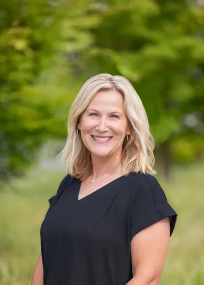 Headshot of Erin Toman, smiling with shoulder-length blonde hair, wearing a black blouse, in front of a blurred green outdoor background.