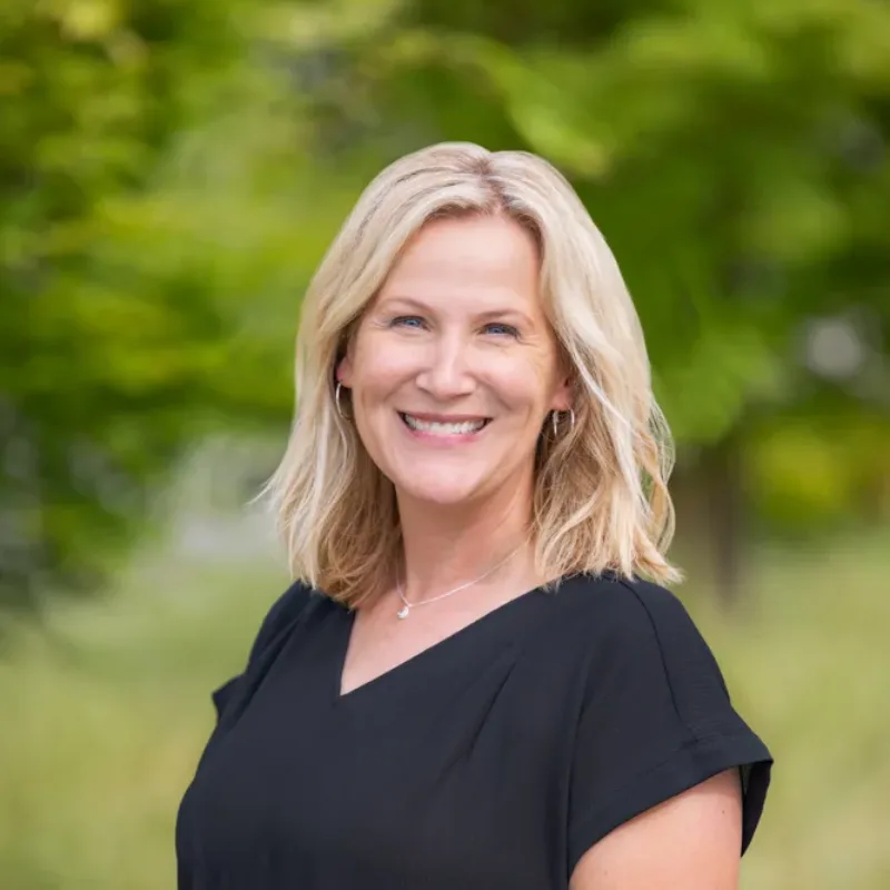 Headshot of Erin Toman, smiling with shoulder-length blonde hair, wearing a black blouse, in front of a blurred green outdoor background.