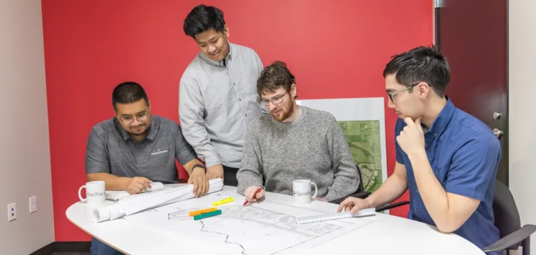 Four MacKay Sposito team members collaborate around a conference table, reviewing site-plan blueprints with scale rulers and highlighters in a red-walled office.