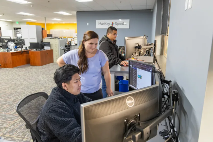 MacKay Sposito team members collaborate at dual-monitor workstations in an open office, with the company logo visible on the wall.