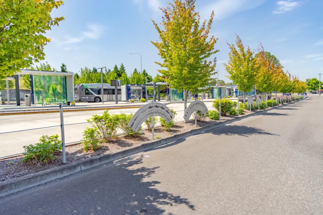 Sunny view of a transit center with glass bus shelters and a bus, bordered by trees and a row of curved metal art panels with fish designs along the sidewalk.