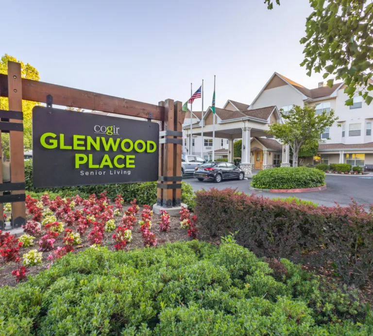 Wooden entrance sign for Cogir Glenwood Place Senior Living amid colorful flowers, with the white multi-story facility, driveway, parked cars, and flagpoles in the background.