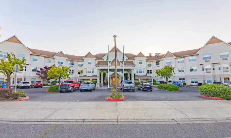 Wide view of a three-story beige building with gabled roofs and a covered entrance (porte-cochère), three flagpoles, and a front parking lot with cars and trimmed shrubs under a clear sky.