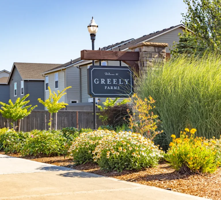 Landscaped neighborhood entrance sign reading “Welcome to Greely Farms” beside ornamental grasses and flowers, with suburban homes and a lamppost under a clear blue sky.