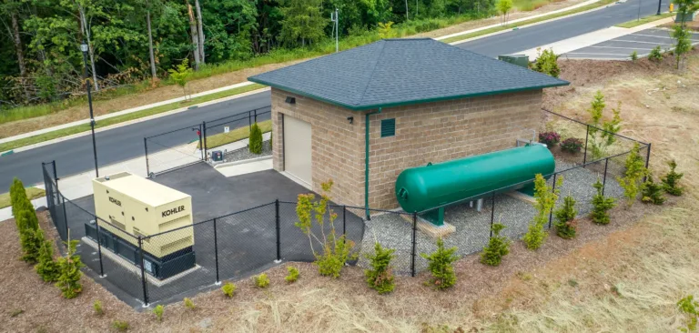 Aerial view of a fenced utility site with a small brick building, a large green horizontal storage tank, and a KOHLER backup generator beside a newly paved road with landscaping.