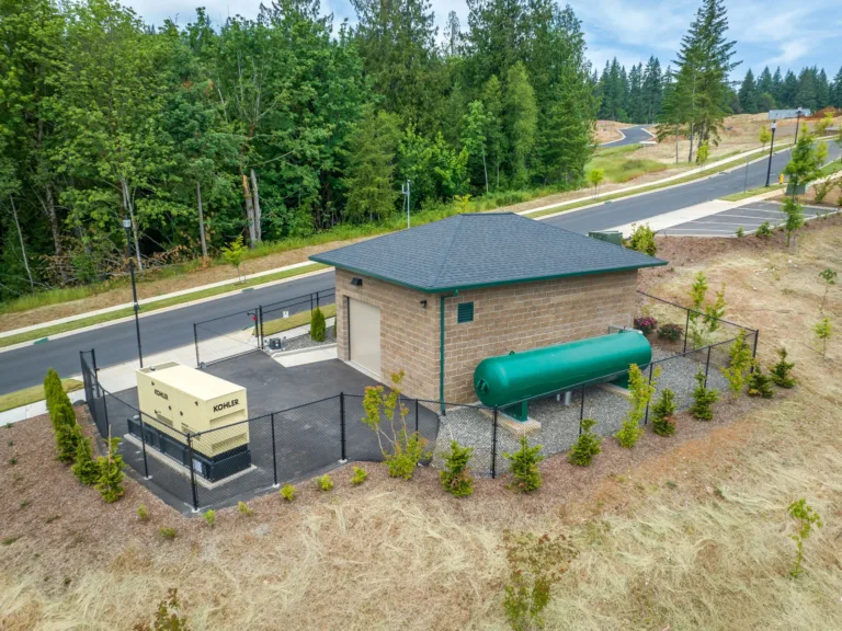 Aerial view of a fenced utility site with a small brick building, a large green horizontal storage tank, and a KOHLER backup generator beside a newly paved road with landscaping.