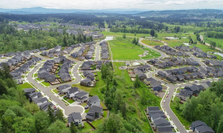 Aerial view of a sprawling suburban neighborhood with curving streets and rows of similar dark‑roofed houses, bordered by green belts, parks, and ponds amid forested hills.