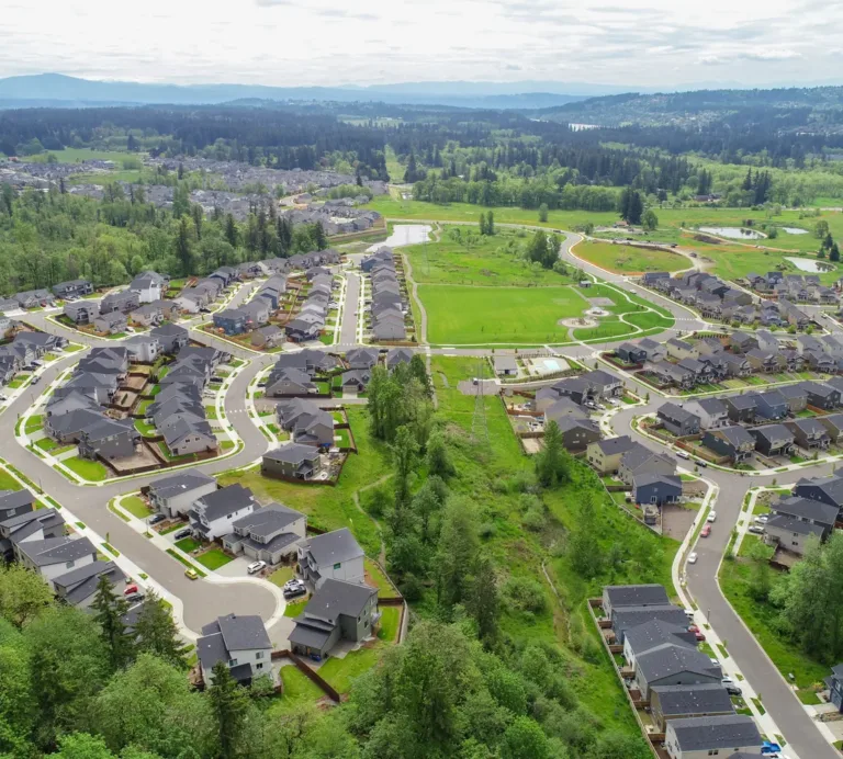 Aerial view of a sprawling suburban neighborhood with curving streets and rows of similar dark‑roofed houses, bordered by green belts, parks, and ponds amid forested hills.