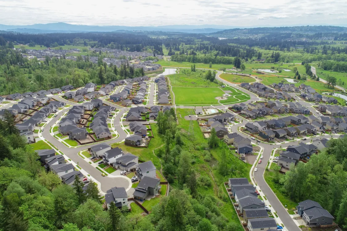 Aerial view of a sprawling suburban neighborhood with winding streets and cul-de-sacs, rows of dark-roofed houses, and a large central green space with paths and ponds, bordered by lush forested hills.