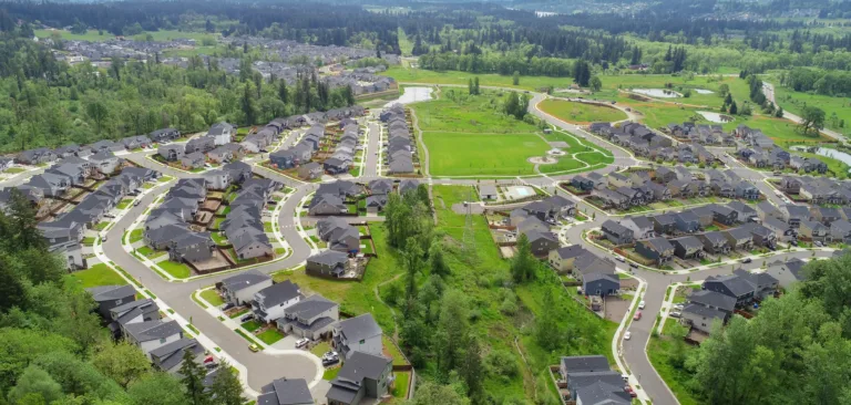 Aerial view of a sprawling suburban neighborhood with curving streets and rows of dark-roofed houses, centered around a large grassy park and small ponds, surrounded by forested hills under a cloudy sky.