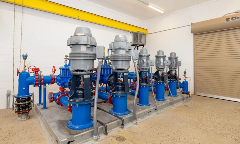 Row of blue industrial pumps with gray vertical motors and red valve wheels mounted on a skid in a clean pump room, with piping, a yellow overhead beam, and a beige roll-up door in the background.