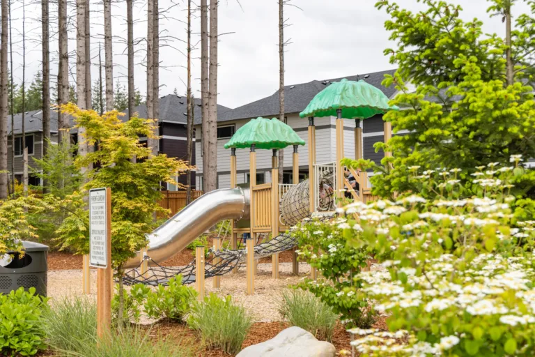 Community playground with green canopy-topped structures, a metal tube slide, and rope bridges, surrounded by landscaped greenery with townhomes and tall trees in the background.
